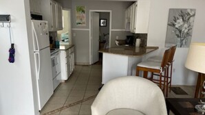 Una cocina con armarios blancos, un refrigerador y una zona de comedor con una silla y una mesa.
