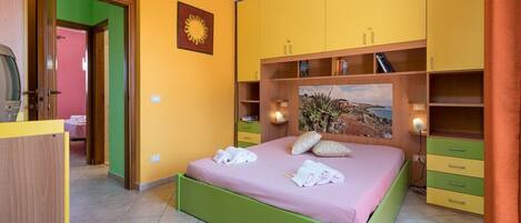 Un dormitorio con una cama verde, armarios amarillos y verdes, un cuadro sobre la cama y una puerta que da a otra habitación.