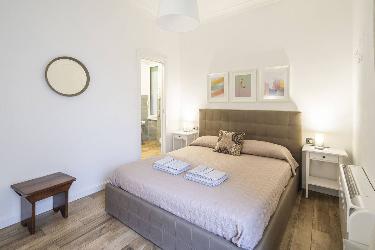Un dormitorio con cama, mesitas de noche, un espejo y cuadros en la pared.
