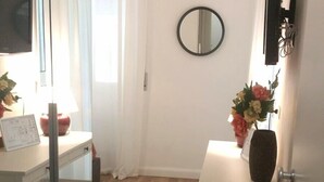 Una habitación con un escritorio blanco, un espejo, un televisor y una pequeña mesa con un jarrón de flores.
