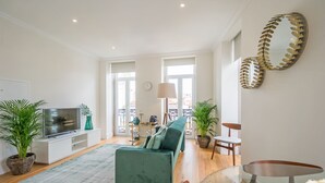 A spacious living room with a green sofa, a TV on a white stand, a dining table with chairs, and a large mirror on the wall.