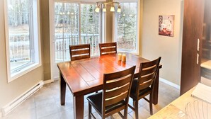 Un coin repas avec une table et des chaises en bois, de grandes fenêtres donnant sur la verdure et un luminaire suspendu.