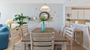 Un comedor con una mesa de madera, sillas blancas, un florero azul verdoso con plantas y una lámpara colgante.