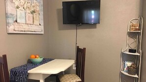 Un comedor con una mesa, sillas, un bol de fruta, un televisor montado en la pared y un estante metálico.