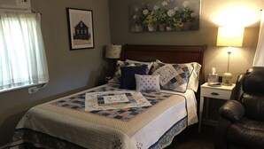 Un dormitorio con cama, mesita de noche, una silla, ventana con cortinas, un reloj de pared, un cuadro enmarcado y una lámpara de pared.