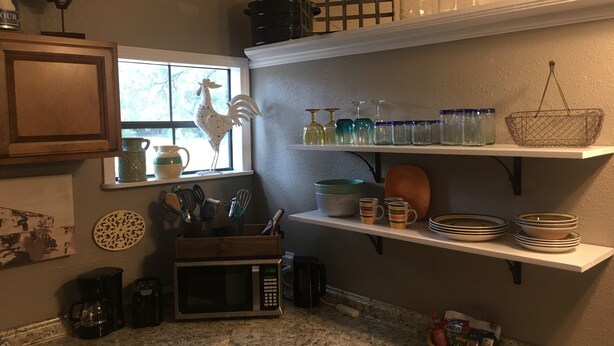 Una cocina con microondas, cafetera y varios utensilios sobre la encimera. Hay estantes arriba con frascos, platos y una cesta.