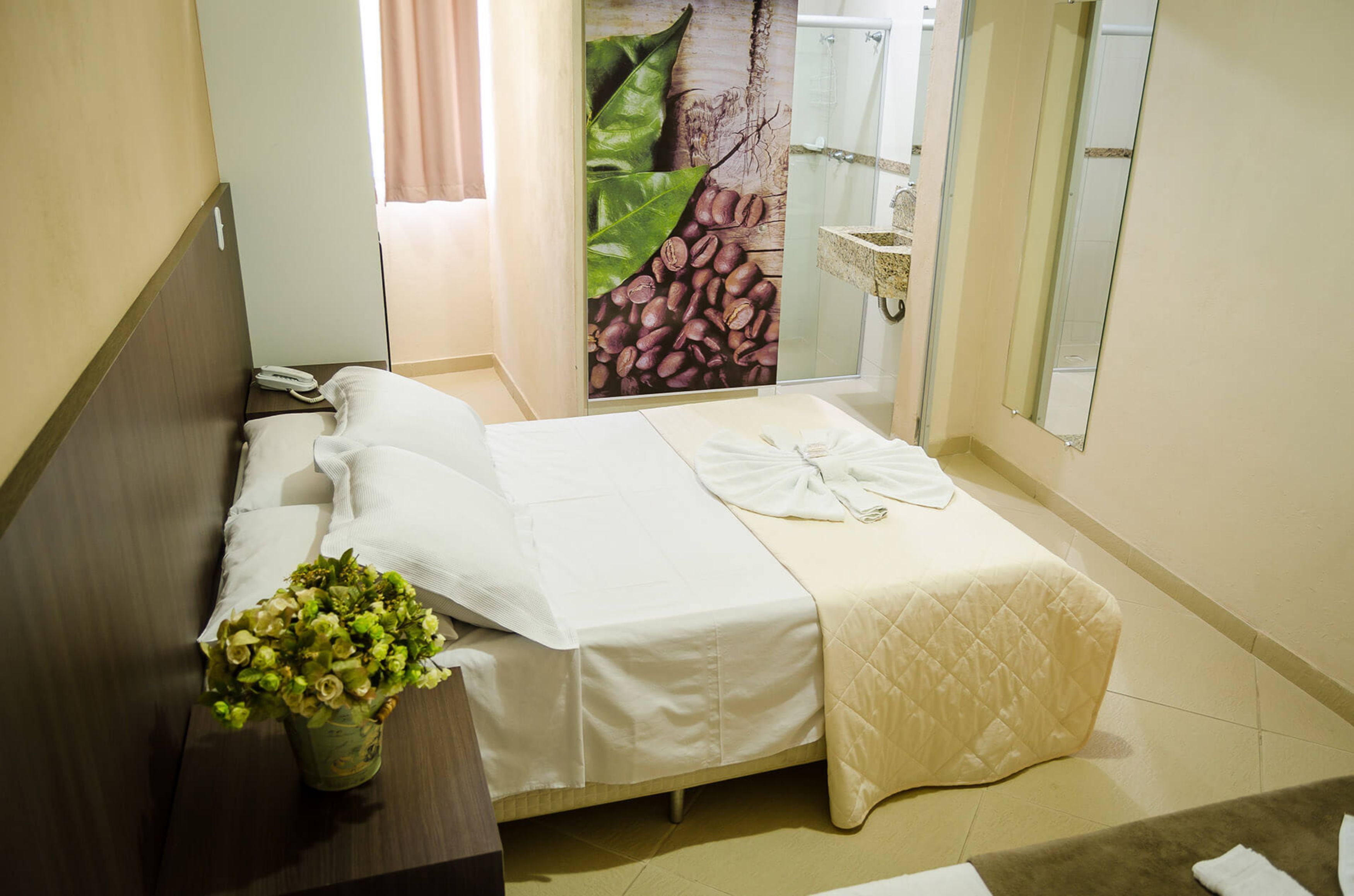A hotel room with a bed, a bedside phone, a vase with flowers, and a large mirror.