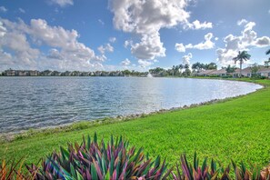 A waterfront property with a grassy area, a body of water, and a row of buildings in the background. - Lake View Villa with a 1 Car Garage, 2 Bedrooms in Lakeside of Naples, Fl (Naples)