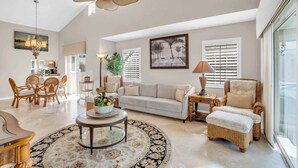 A spacious living room with a large ceiling fan, a dining area with wicker furniture, and a view of the outdoors through large windows. - Lake View Villa with a 1 Car Garage, 2 Bedrooms in Lakeside of Naples, Fl (Naples)