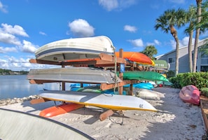 A row of paddleboards stacked on a wooden stand by a sandy beach with a body of water and palm trees in the background. - Lake View Villa with a 1 Car Garage, 2 Bedrooms in Lakeside of Naples, Fl (Naples)