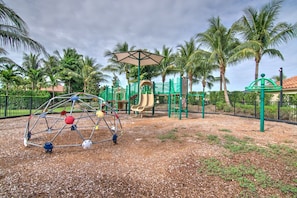 A playground with a climbing dome, slides, and a covered play area, surrounded by palm trees and a fence.