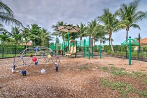 A playground with a climbing dome, slides, and swings, surrounded by palm trees and a fence.