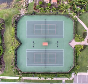 An aerial view of a tennis court complex with two courts, surrounded by landscaped areas and palm trees.