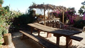 Un espacio de descanso rústico al aire libre con una mesa y bancos de madera, rodeado de vegetación y una estructura de techo de paja.