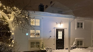 A snow-covered house with a black door, a small Christmas tree, and string lights.