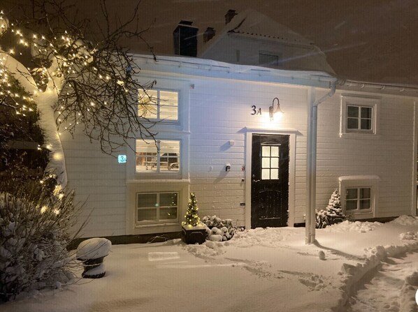 Una casa cubierta de nieve con una puerta negra, un pequeño árbol de Navidad y luces de adorno.