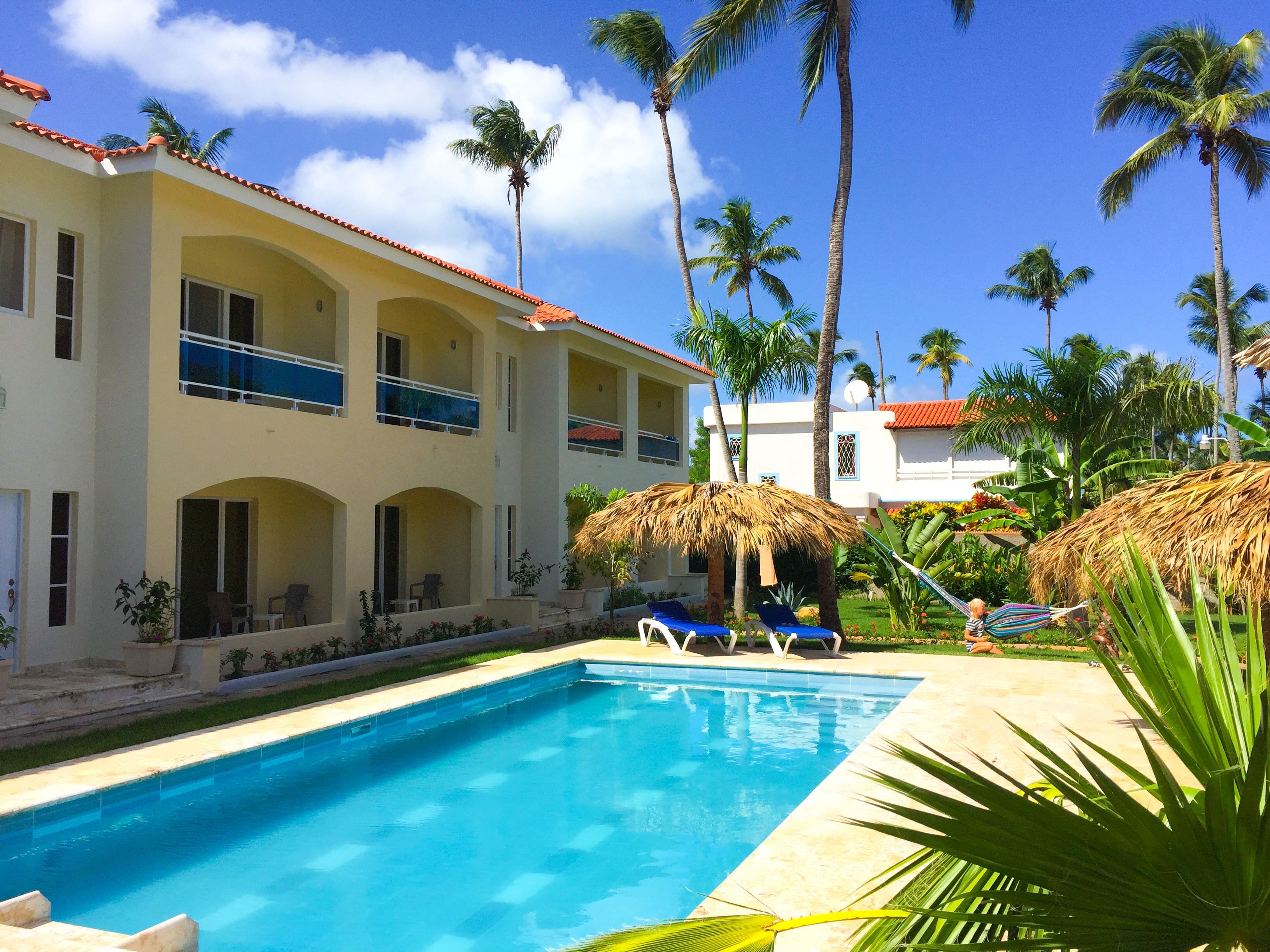 A hotel with a swimming pool, palm trees, and a thatched-roof structure.