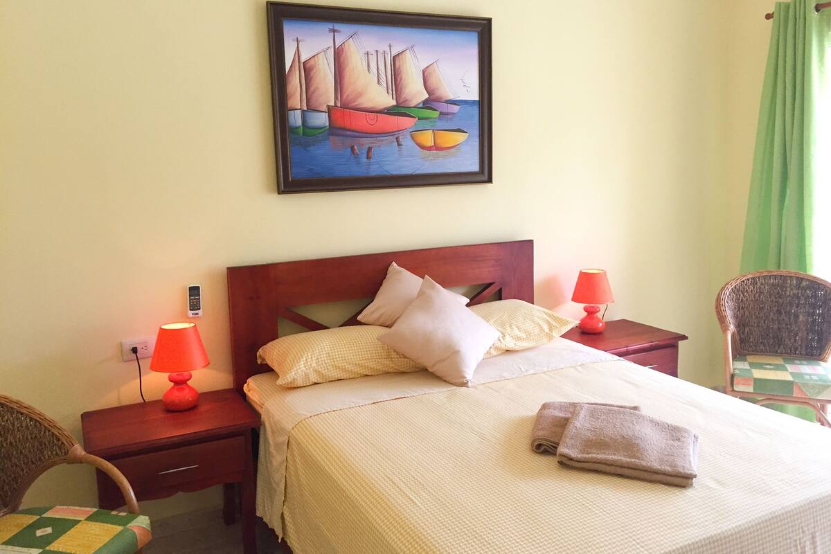 A neatly made bed with pillows and a folded towel, a painting of sailboats on the wall, and a chair with a checkered cushion.