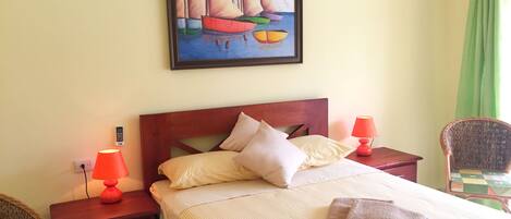 Un lit bien fait, avec des oreillers et une serviette pliée, un tableau représentant des voiliers accroché au mur, et une chaise avec un coussin à carreaux.