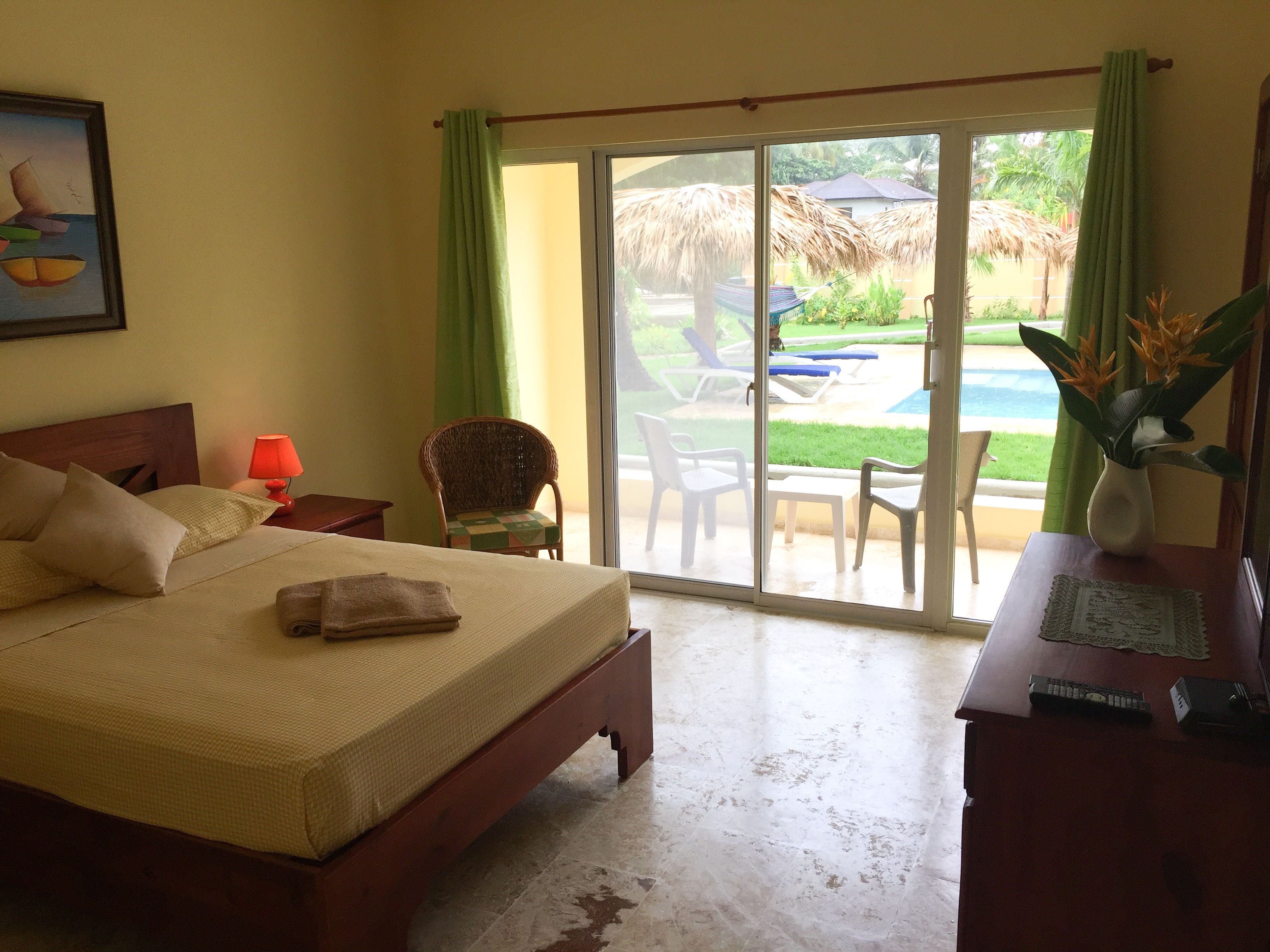 A hotel room with a bed, a chair, a table, a lamp, and a view of an outdoor pool area.