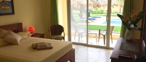 A hotel room with a bed, a chair, a table, a lamp, and a view of an outdoor pool area.