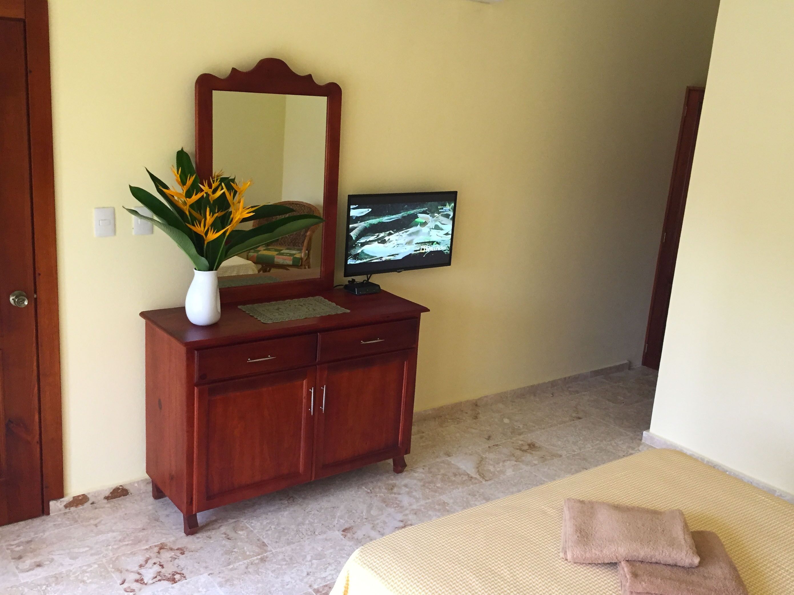 Un dormitorio con un aparador de madera, un espejo, un televisor y una cama con una colcha color beige y dos almohadas.