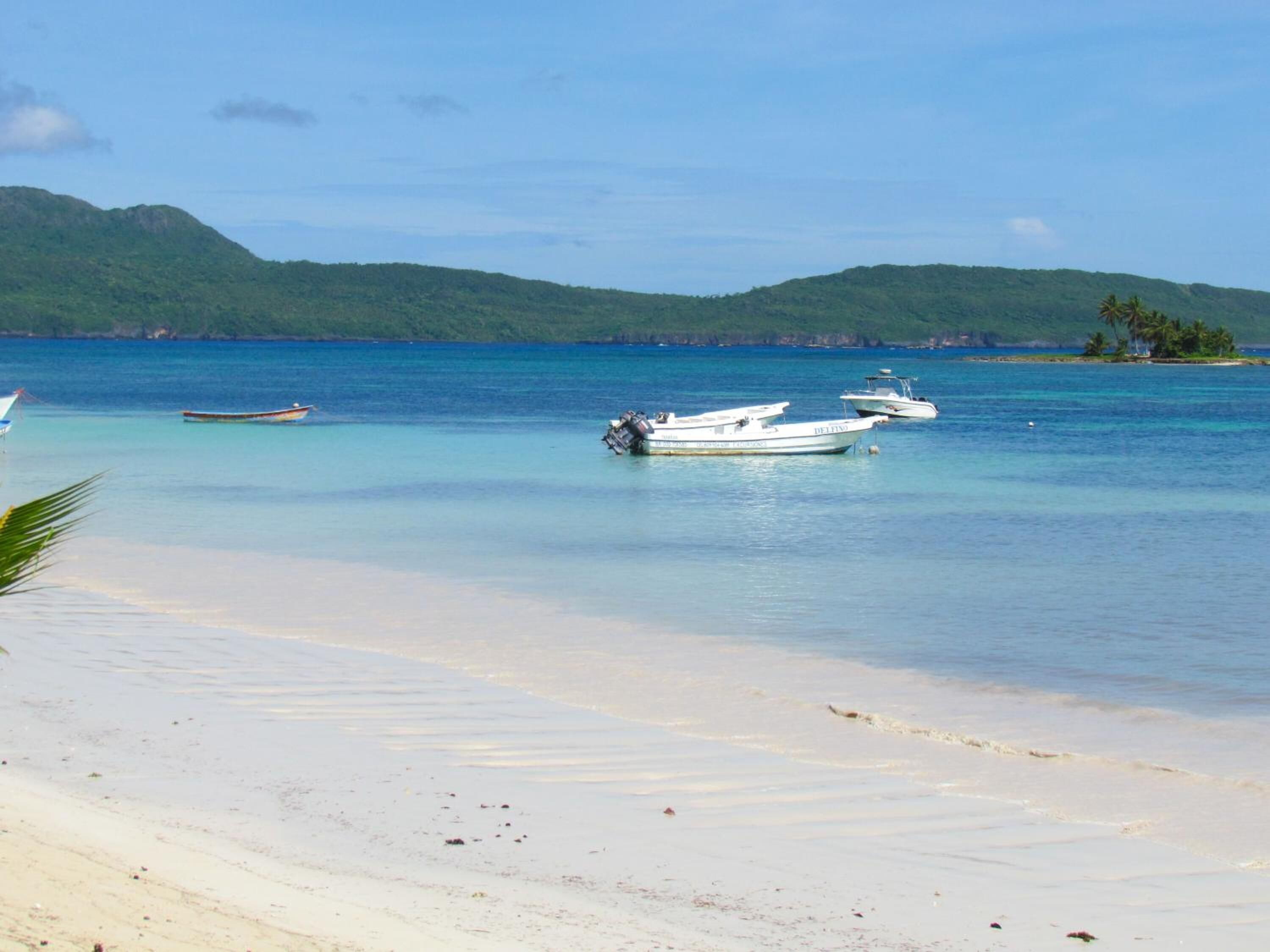 A beach with several boats anchored in calm waters, surrounded by lush green hills.