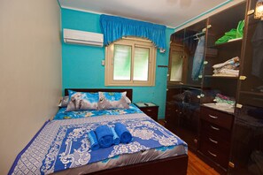 A bedroom with a bed, a window with blinds, a wardrobe, and an air conditioning unit.