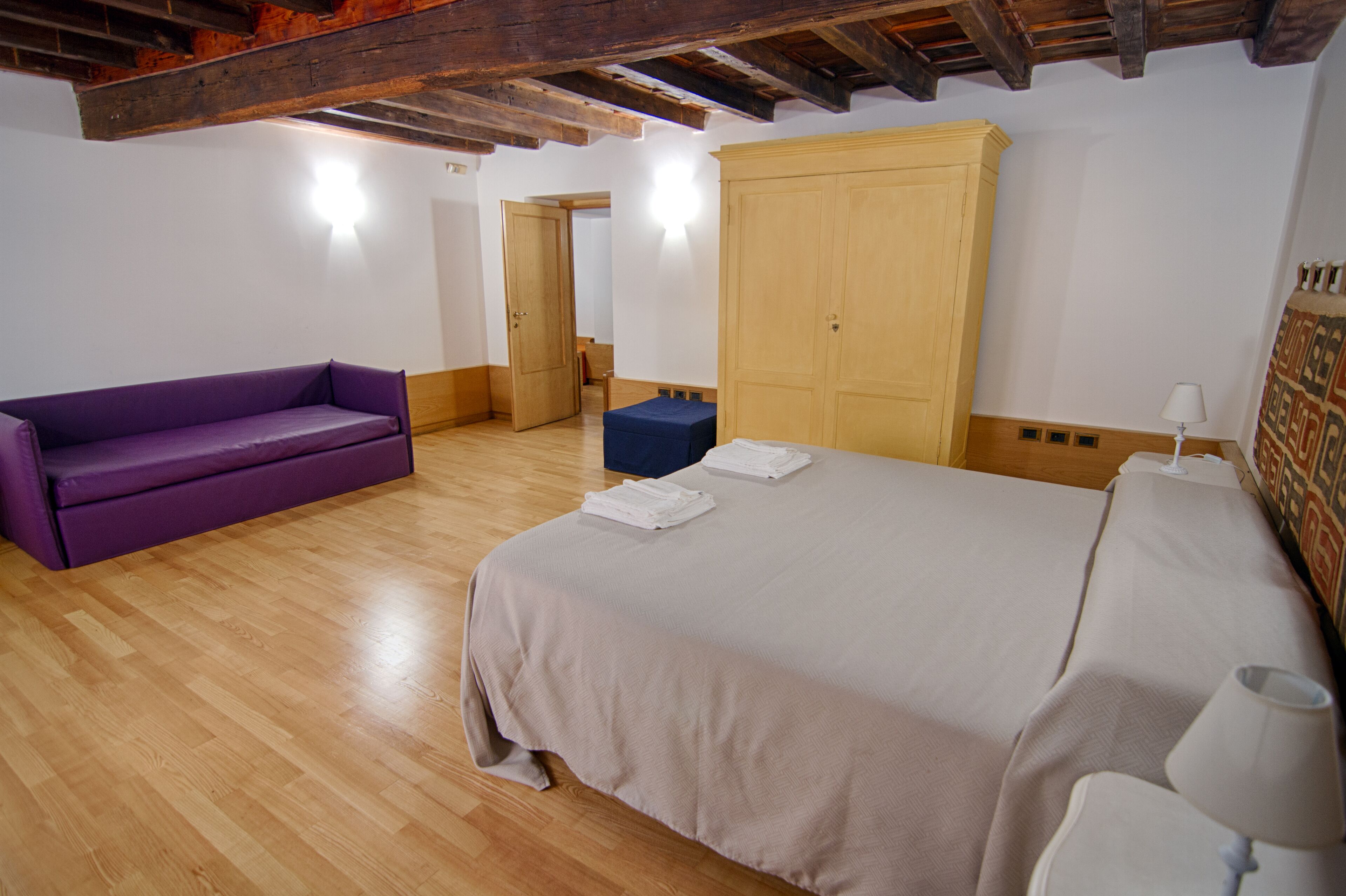 A room with a bed, a purple sofa, a wooden wardrobe, and a small blue ottoman.