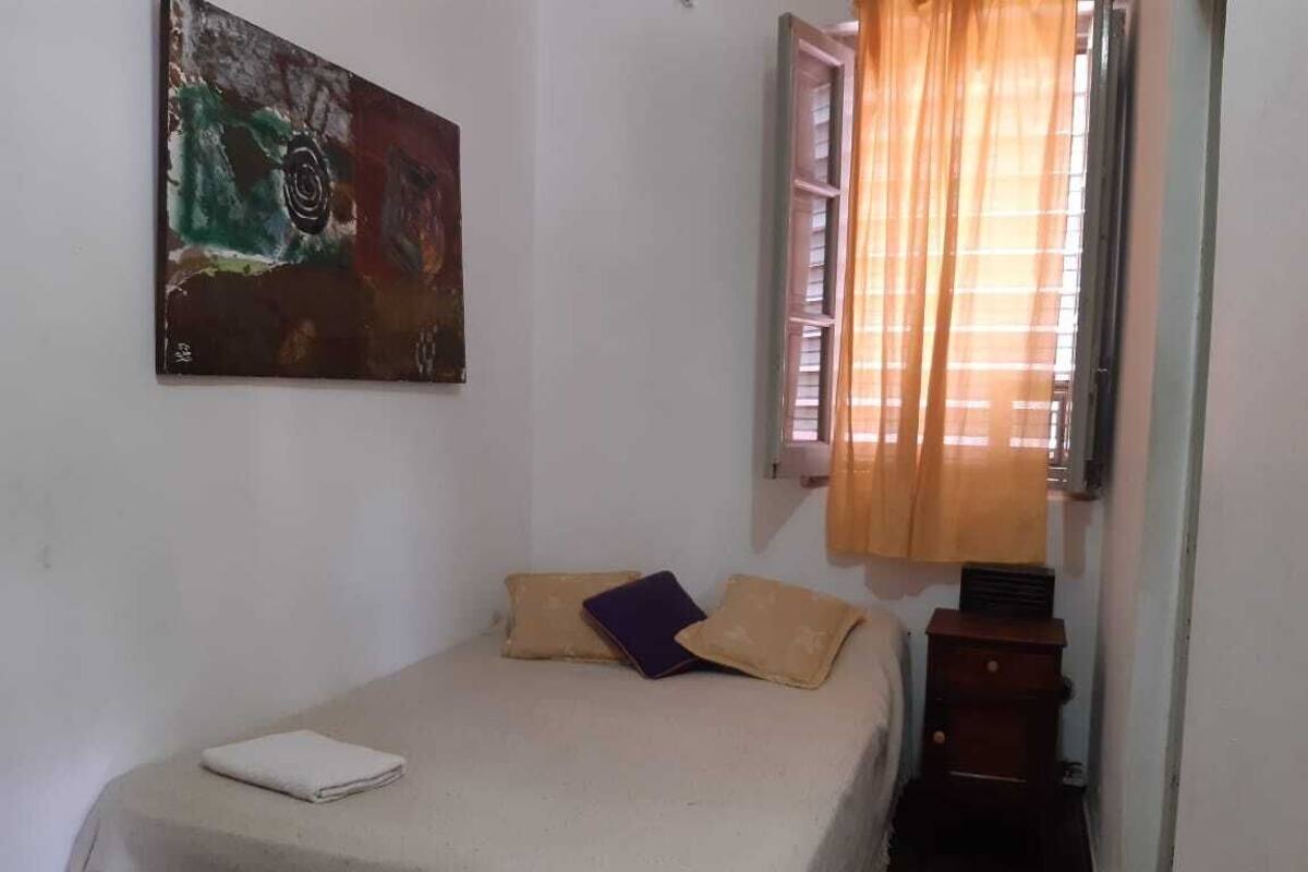 Un dormitorio con cama, mesita de noche, ventana con cortinas y un cuadro en la pared.