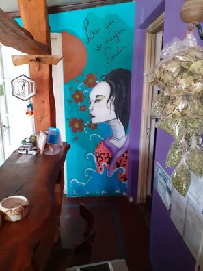 A room with a mural, a wooden table, and a plastic bag of dried leaves. - Hostel El Viaje (Mina Clavero)