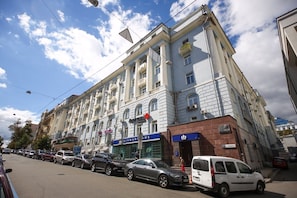 A multi-story building with a classical architectural style, featuring a prominent entrance with a sign, a visible bank, and a parking area with cars.