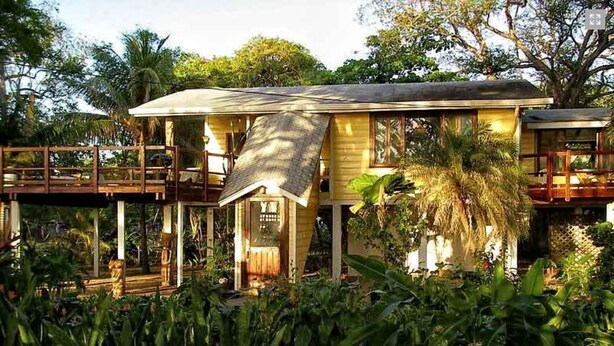 Una casa de dos pisos con un porche cubierto y una terraza de madera, rodeada de exuberante vegetación tropical.
