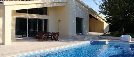 A modern house with a swimming pool, a patio with outdoor furniture, and a large window overlooking the pool.