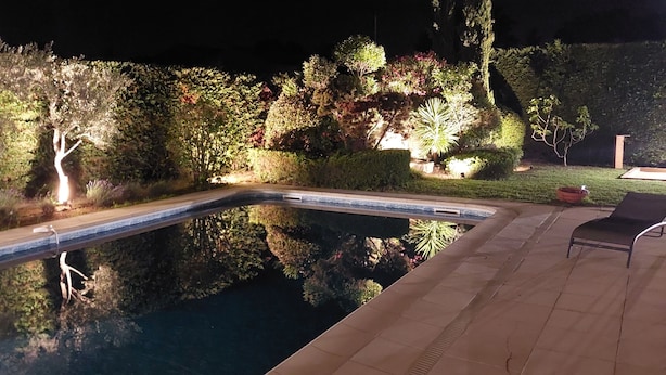 Une piscine à bord incurvé, entourée de haies et d’arbres bien entretenus, et un espace patio avec une chaise.