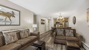 A living room with two sofas, a coffee table, and a rug. There is a kitchen in the background with a refrigerator and a dining area.