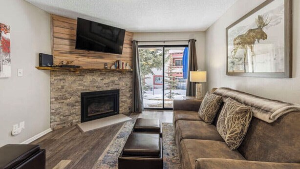A living room with a stone fireplace, a flat-screen TV mounted on a wooden wall, a brown sofa, and a large window with a view of the outside.