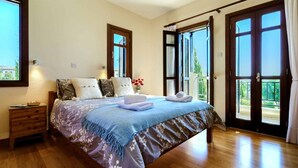 A bedroom with a bed, wooden nightstand, and a view through open French doors to a balcony. - Apartment A12 - Nesoi (Kouklia)