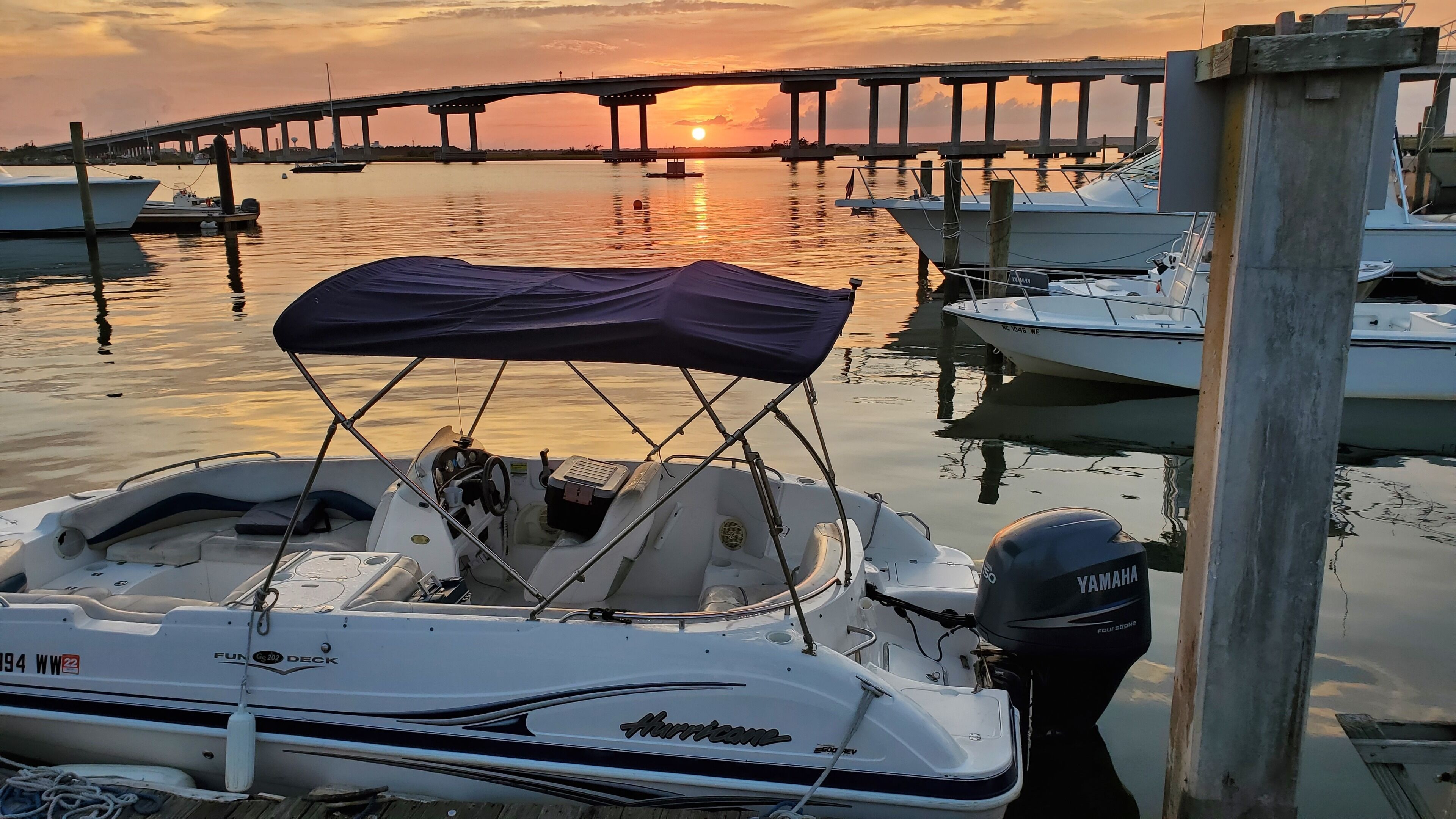 Een jachthaven met aangemeerde boten, een brug op de achtergrond en een zonsondergang.