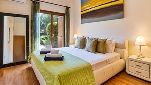 A hotel room with a large bed, a nightstand with a lamp, a view of greenery outside, and a painting on the wall.
