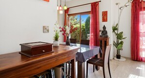 A dining area with a wooden table, chairs, a vase with flowers, a Buddha statue, and a view of greenery through the window.