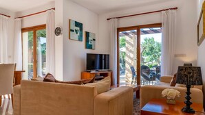 A living room with beige sofas, a TV, a table with a lamp, and a view of the outside. - Villa HG24 Cardia - Three Bedroom Villa, Sleeps 6 (Kouklia)