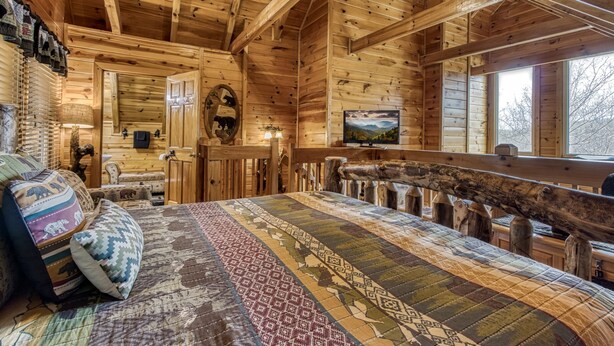 A cozy log cabin room with a large bed, wooden walls, a ceiling fan, and a window with a view of trees. - Located 1.5 mi. from the Parkway in Pigeon Forge. (Pigeon Forge)
