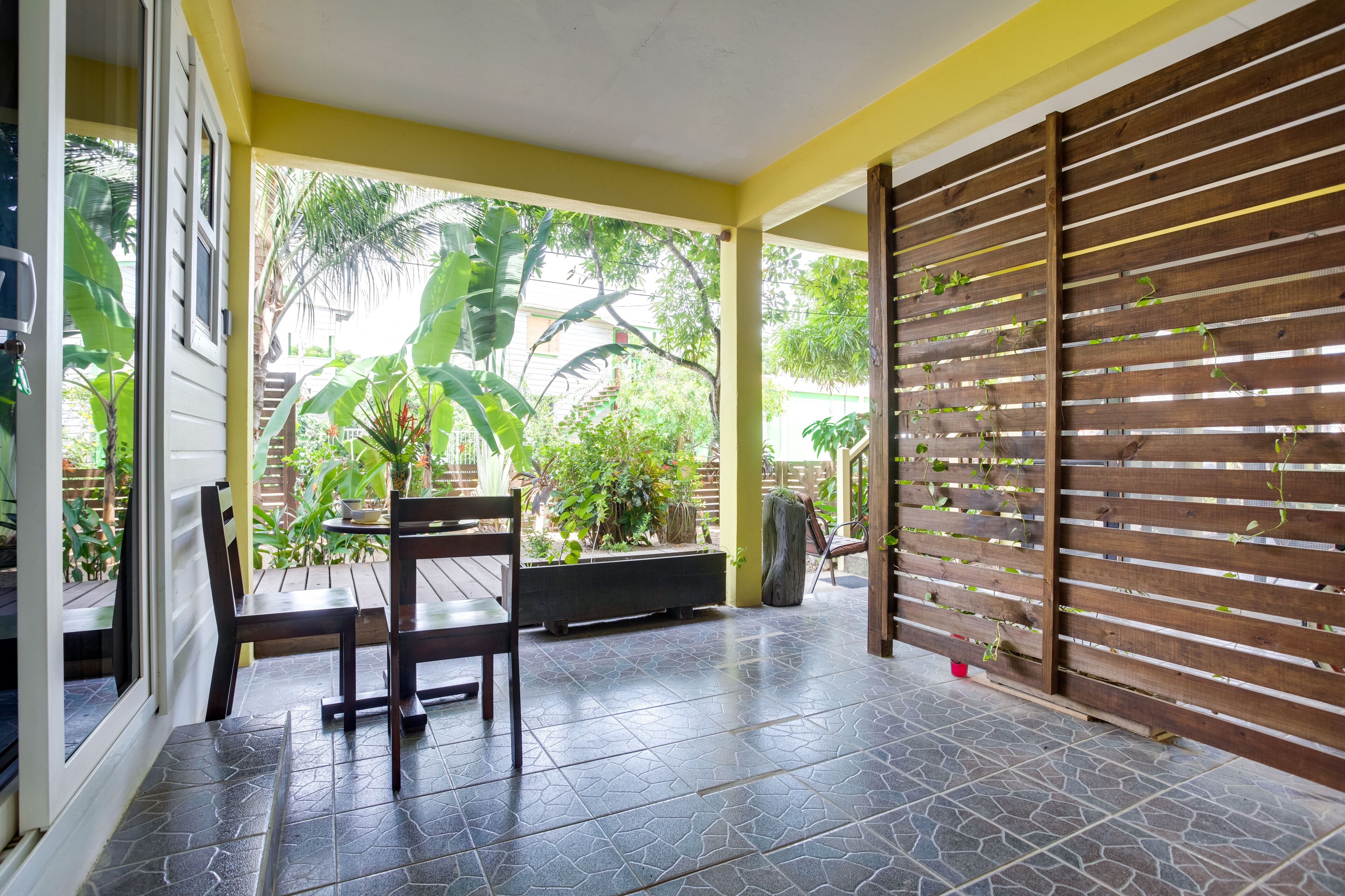 Un patio cubierto con un banco y sillas de madera, piso de baldosas y vistas al exterior con vegetación.