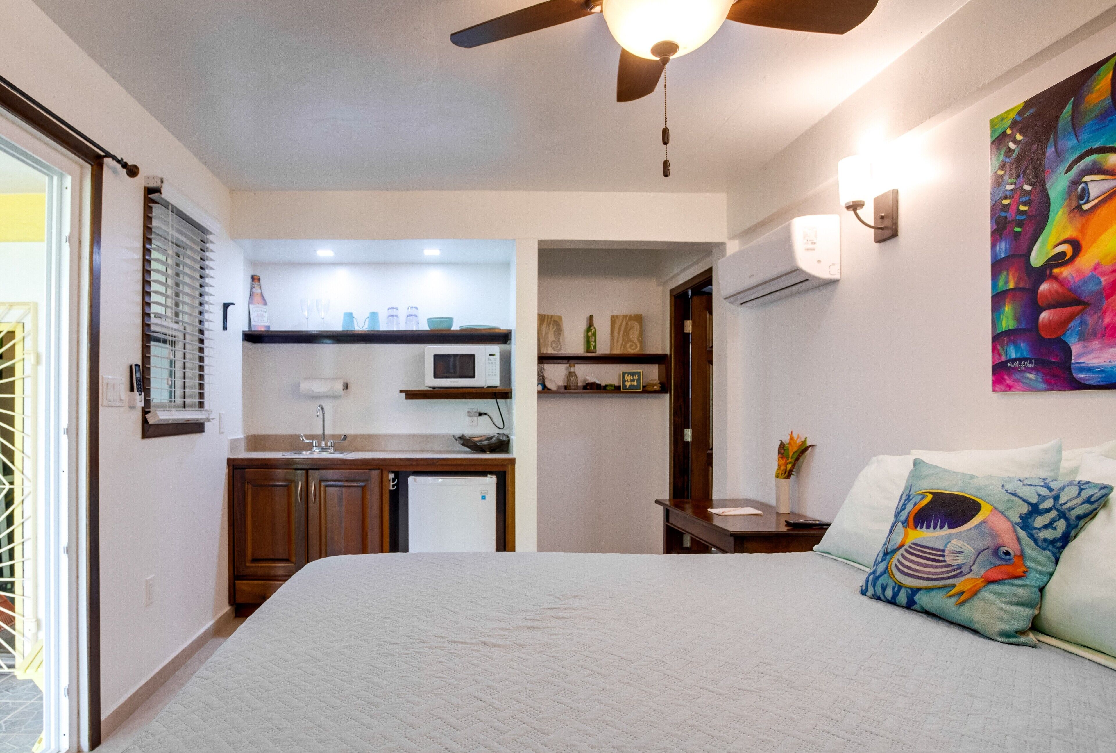 Un dormitorio con cama, mesita de noche, almohada con diseño de pez, ventilador de techo y vista a la cocina contigua.