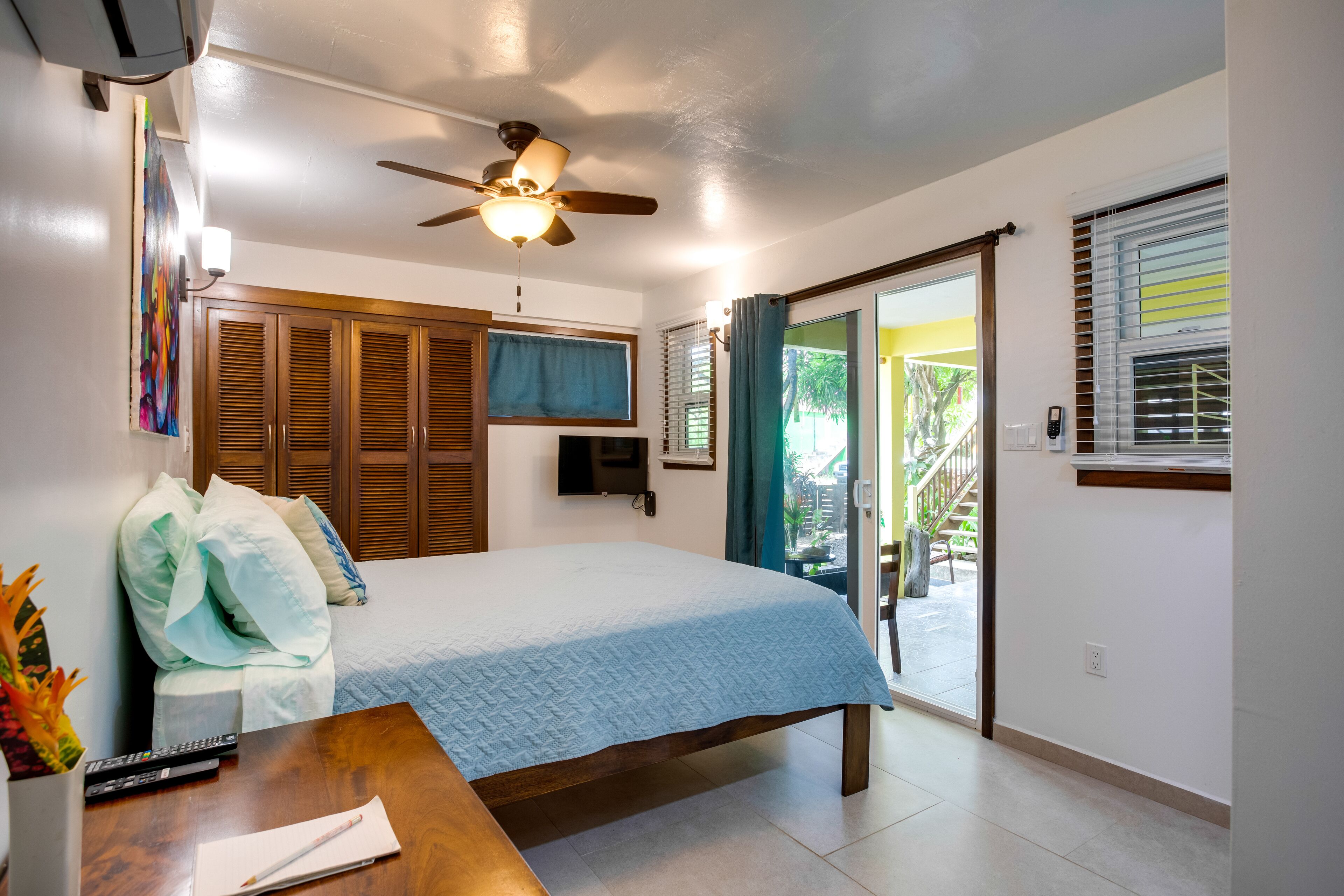 A bedroom with a bed, wooden wardrobe, ceiling fan, and a view to the outside.