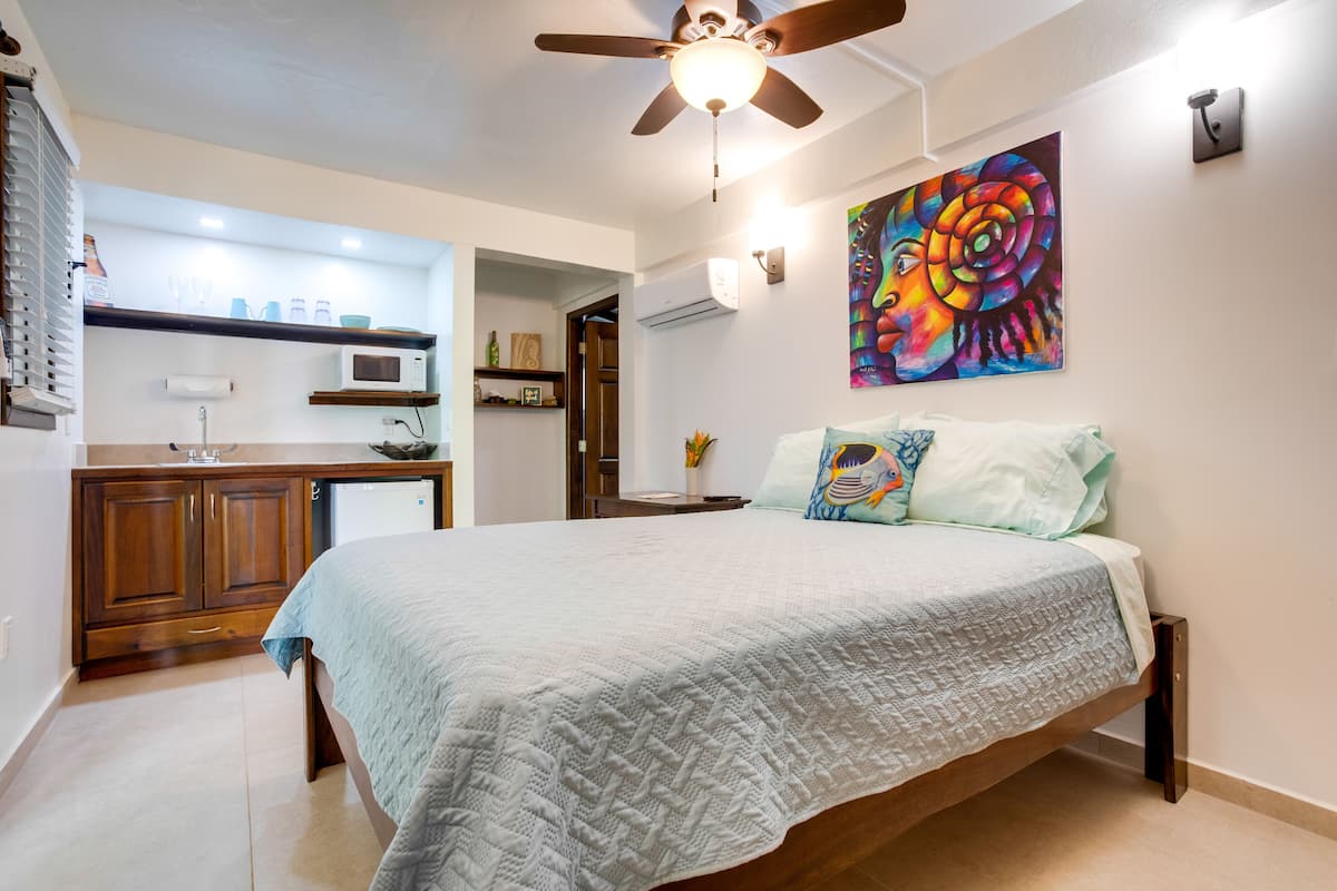 A bedroom with a bed, a ceiling fan, a painting, and a kitchenette in the background.