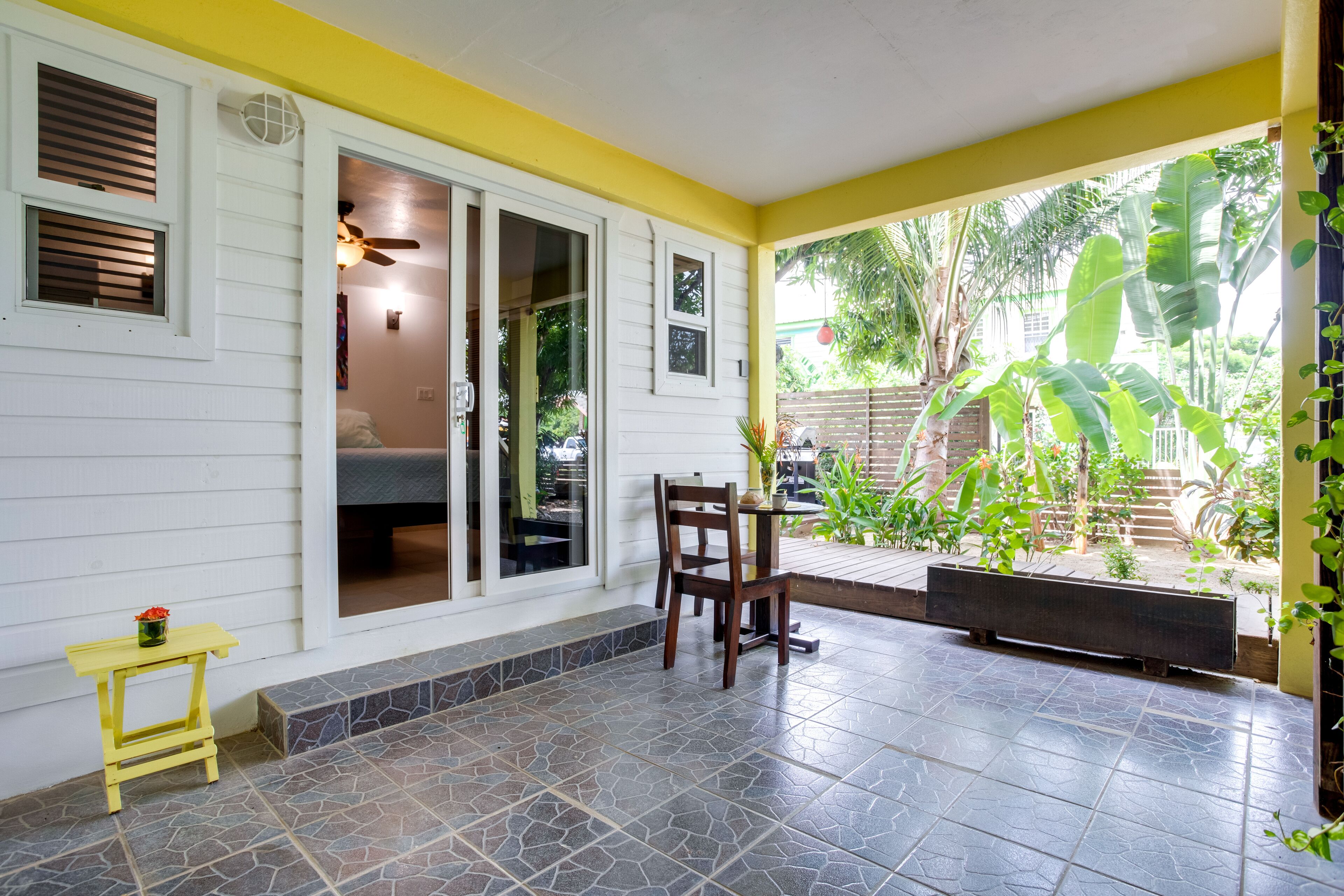 A covered patio with a table and chair, a small yellow table, and a view of the garden.