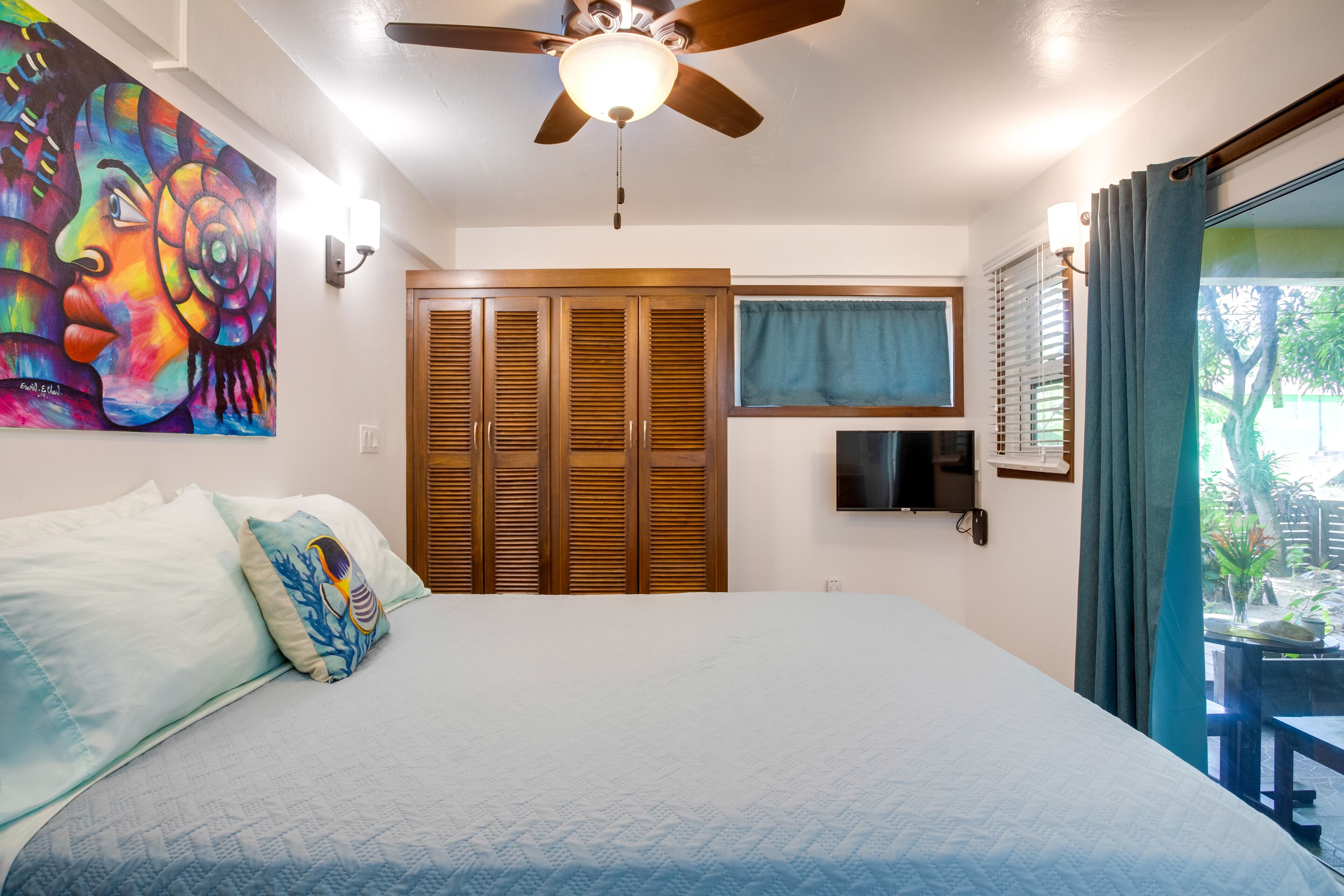 A bedroom with a bed, a ceiling fan, a TV, and a view of the outdoors.