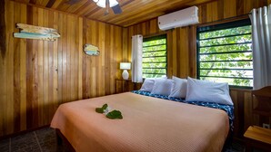 Una habitación de madera con una cama, un ventilador de techo, un equipo de aire acondicionado, una ventana con cortinas y un cartel en la pared.